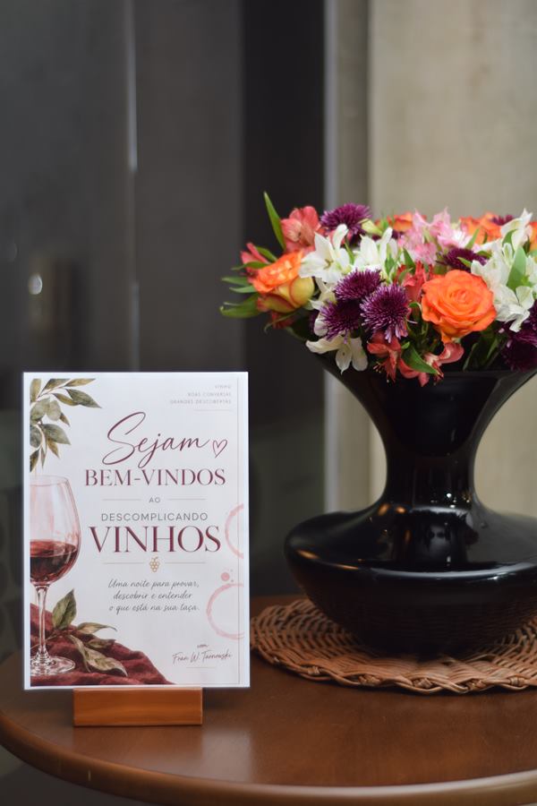 Placa de boas-vindas ao Descomplicando Vinhos, por Franciele Tarnwski, ao lado de um arranjo de flores